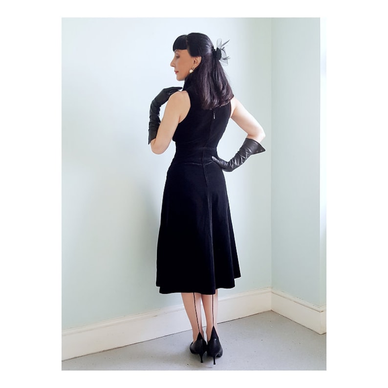 May include: A black velvet dress with a sleeveless bodice and a fitted waist. The dress has a flared skirt and a high neckline. The model is wearing black gloves and black heels.