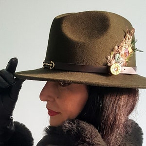 May include: Olive green fedora hat with a brown leather band and gold-colored buckle. The hat is decorated with a cluster of colorful feathers and a gold-colored bullet casing. The brim is wide and the hat is being held by a person wearing black gloves.