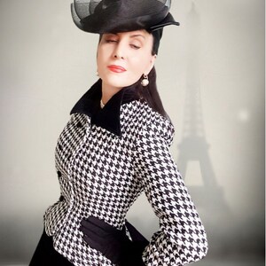 May include: A woman wearing a black and white houndstooth jacket with a black velvet collar and a black hat with a veil. She is standing in front of a blurred image of the Eiffel Tower.