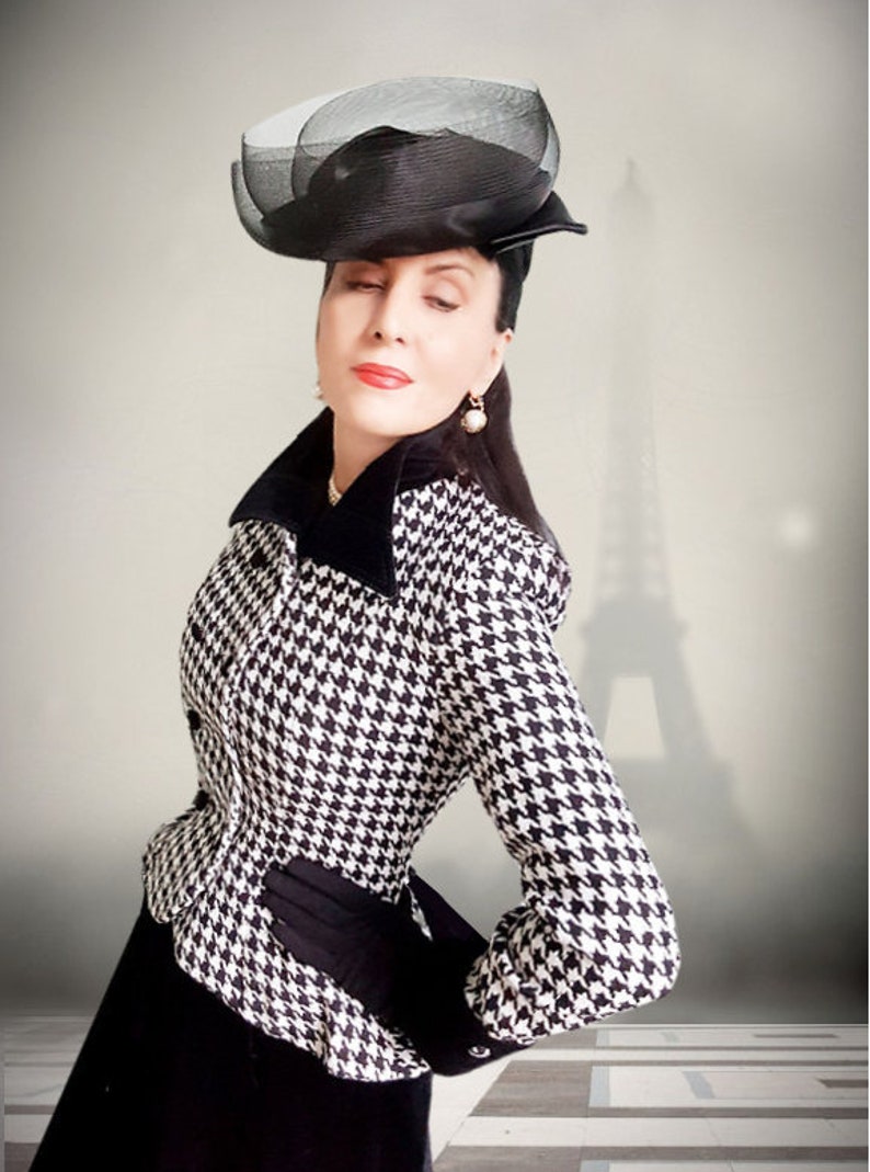 May include: A woman wearing a black and white houndstooth jacket with a black velvet collar and a black hat with a veil. She is standing in front of a blurred image of the Eiffel Tower.