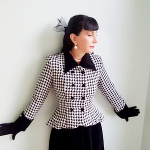 May include: A black and white houndstooth jacket with a black velvet collar and cuffs. The jacket has a fitted silhouette and a double-breasted closure. The model is wearing black gloves and a pearl necklace.