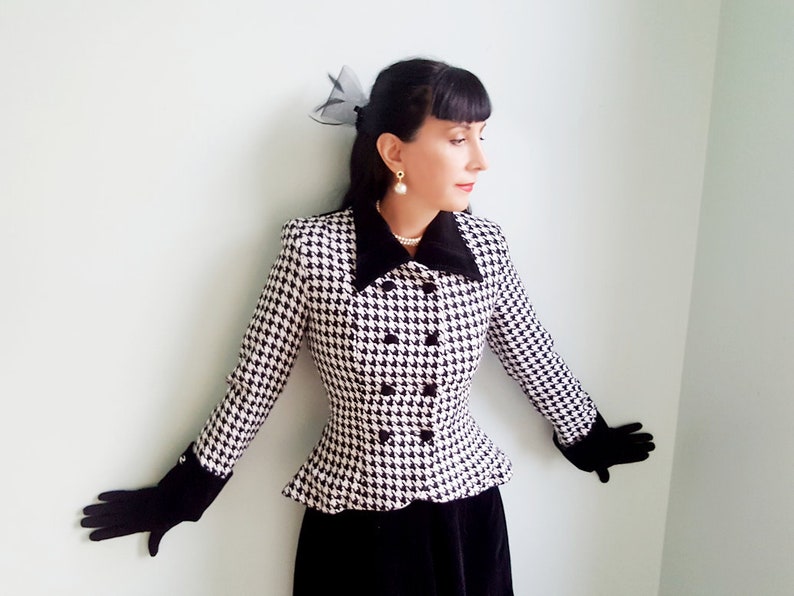 May include: A black and white houndstooth jacket with a black velvet collar and cuffs. The jacket has a fitted silhouette and a double-breasted closure. The model is wearing black gloves and a pearl necklace.