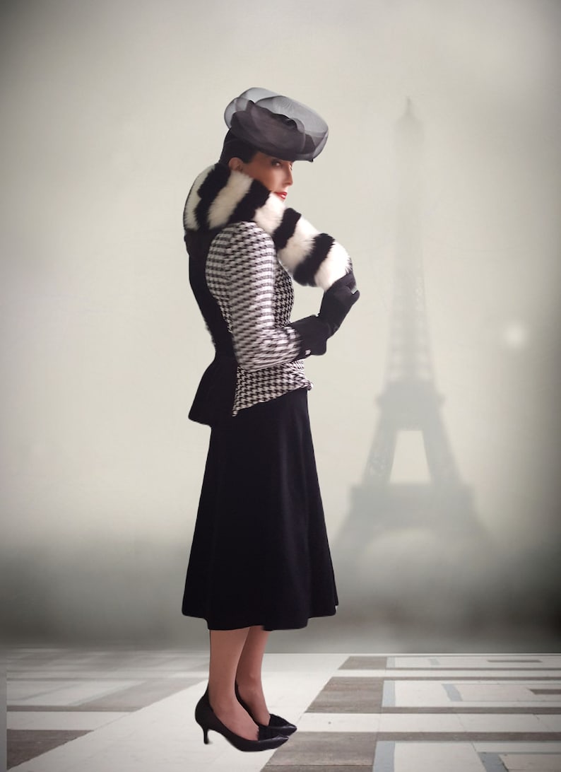 May include: A woman wearing a black and white houndstooth jacket with a black and white fur collar and a black hat. She is standing in front of a blurry image of the Eiffel Tower.