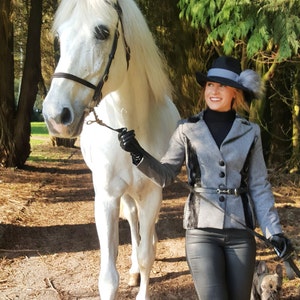 Puede incluir: Una mujer con una chaqueta gris y pantalones negros camina con un caballo blanco y un perro pequeño marrón y blanco. La mujer lleva un sombrero negro con un pompón blanco.