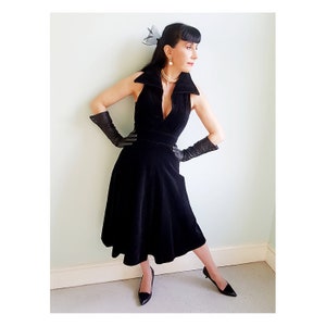 May include: A woman wearing a black velvet dress with a plunging neckline and a high collar. She is wearing black gloves and black heels. She has her hair styled in a black bob with a black bow.