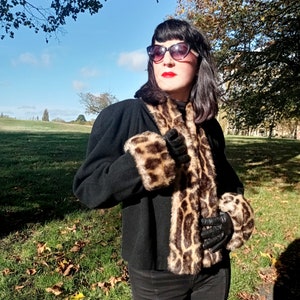 Pode incluir: Uma mulher vestindo uma jaqueta preta com gola e punhos de pele de leopardo. Ela está usando óculos de sol pretos e batom vermelho. O fundo é um campo gramado verde com árvores ao longe.