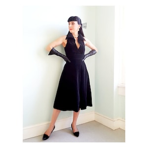 May include: A black velvet dress with a deep V-neckline and a fitted bodice. The dress has a full skirt and is sleeveless. The model is wearing black leather gloves and black heels.
