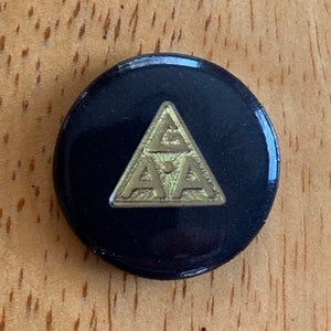 May include: A black button with a gold triangle featuring the letters "AA" inside. The button is on a wooden surface.