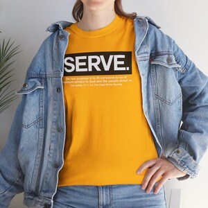 Peut inclure: T-shirt jaune moutarde avec le mot « SERVE » dans un rectangle noir. En dessous, une citation sur le service. La personne porte une veste en jean bleu clair et un jean bleu.