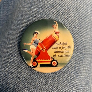 May include: A round button with a vintage illustration of two women in a red cannon. The text on the button reads "rocketed into a fourth dimension of existence".