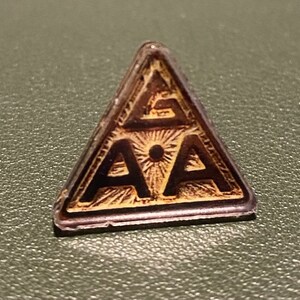 May include: A gold-colored triangular pin with the letters "AA" inside a circle with a dot in the center. The letters are surrounded by a sunburst design.