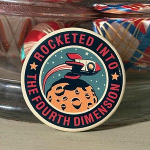 May include: A round, white button with a blue, red, and white design. The button features a rocket ship flying through space with a planet in the background. The text "ROCKETED INTO THE FOURTH DIMENSION" is written around the edge of the button.