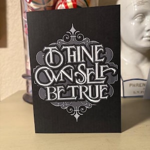 May include: A black card with the text "THINE OWN SELF BE TRUE" in a decorative, vintage-style font. The text is in white with a gray shadow, and the card has ornate flourishes.