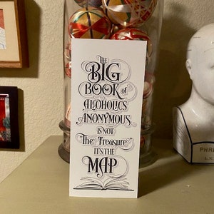 May include: A white bookmark with black text that reads "The Big Book of Alcoholics Anonymous is not "The Treasure" it's the Map". The bookmark features an open book illustration.