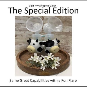 K&ouml;nnte beinhalten: The Special Edition Milchpumpe mit Kuh-Akzenten. Die Pumpe hat transparente Flaschen und steht auf einem Holztablett mit wei&szlig;en Blumen. Der Text "Same Great Capabilities with a Fun Flare" ist sichtbar.