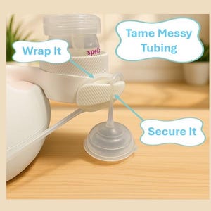 K&ouml;nnte beinhalten: Eine wei&szlig;e und transparente Milchpumpe mit den Texten &bdquo;Wrap It&ldquo;, &bdquo;Tame Messy Tubing&ldquo; und &bdquo;Secure It&ldquo;. Die Pumpe befindet sich auf einer hellbraunen Oberfl&auml;che.