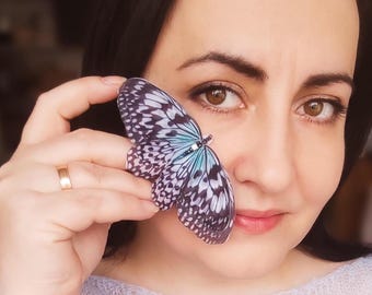 May include: A woman holds a blue and white butterfly brooch near her face. The butterfly has black and white markings and a light blue center.