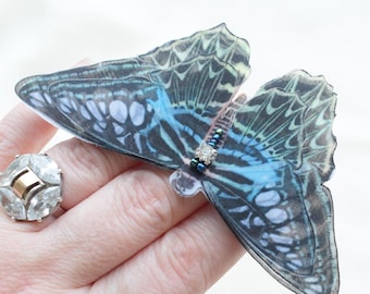 May include: A hand holding a blue and green butterfly brooch with iridescent wings and black bead accents.