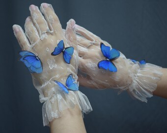 May include: A pair of sheer white lace gloves featuring a delicate floral pattern and ruffled cuffs. Each glove is adorned with multiple vibrant blue butterfly embellishments attached to the fabric, creating a textured and layered appearance.