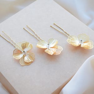 May include: Three gold-colored hair pins with delicate flower designs. Each pin features a detailed, layered petal arrangement and a small, clear gem at the center. The pins are arranged on a light beige surface, with a soft, white fabric in the background.