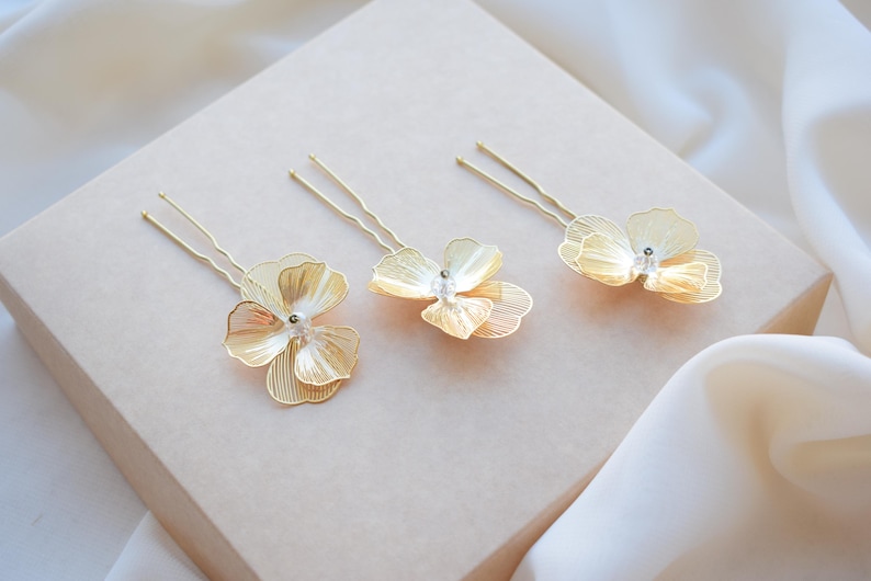 May include: Three gold-colored hair pins with delicate flower designs. Each pin features a detailed, layered petal arrangement and a small, clear gem at the center. The pins are arranged on a light beige surface, with a soft, white fabric in the background.