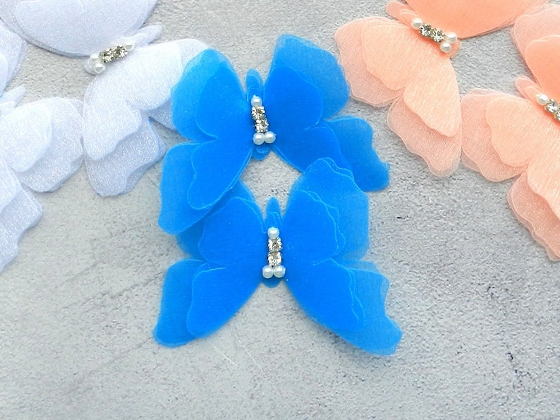 May include: Two blue layered tulle butterfly appliques with rhinestone accents. The butterflies are on a gray background.