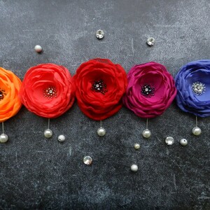 May include: Five fabric flower brooches in shades of orange, red, pink, and purple with a rhinestone center. Each brooch is attached to a silver pin with a pearl dangling from the bottom.