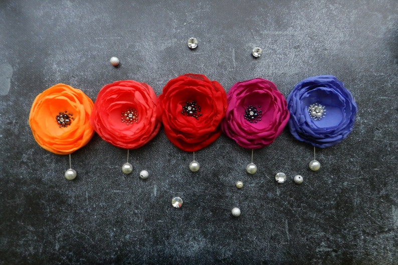 May include: Five fabric flower brooches in shades of orange, red, pink, and purple with a rhinestone center. Each brooch is attached to a silver pin with a pearl dangling from the bottom.
