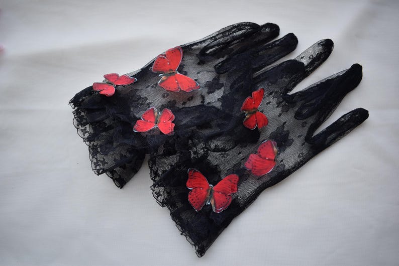 May include: A pair of black lace gloves adorned with vibrant red butterfly appliqu&eacute;s. The gloves have a delicate, floral lace pattern and scalloped edges. The butterflies are evenly spaced across the gloves.