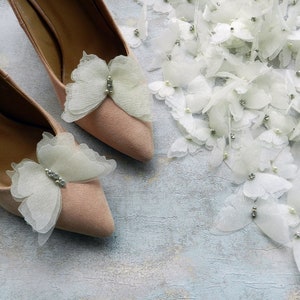 May include: A pair of light pink high heel shoes with delicate white butterfly embellishments. Each butterfly has a small rhinestone in the center.