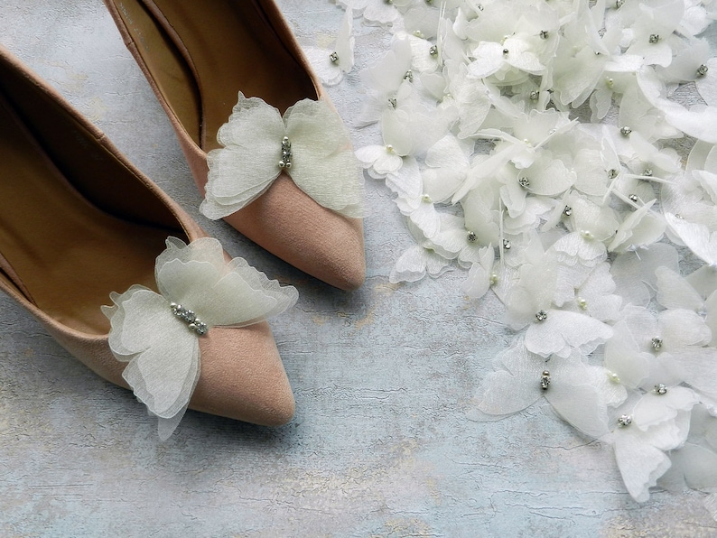 May include: A pair of light pink high heel shoes with delicate white butterfly embellishments. Each butterfly has a small rhinestone in the center.