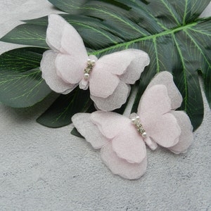 May include: Two delicate pink tulle butterfly hair clips with pearl and rhinestone accents. The clips are on a gray background with a green leaf.