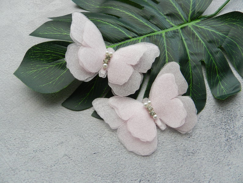 May include: Two delicate pink tulle butterfly hair clips with pearl and rhinestone accents. The clips are on a gray background with a green leaf.