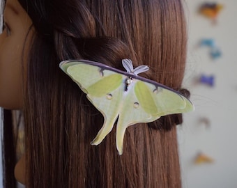May include: A light green and yellow moth hair clip with a silver accent is attached to brown hair. The clip is shaped like a luna moth with detailed wing patterns. The hair accessory adds a touch of nature-inspired elegance.