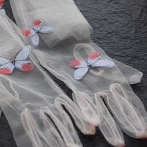May include: A pair of sheer, light pink gloves adorned with delicate butterfly appliqu&eacute;s. The butterflies feature pale blue wings with red accents. The gloves are made of a fine, semi-transparent material, and are set against a dark, textured background.
