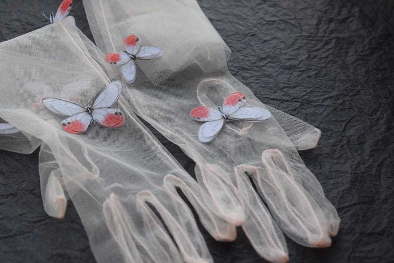 May include: A pair of sheer, light pink gloves adorned with delicate butterfly appliqu&eacute;s. The butterflies feature pale blue wings with red accents. The gloves are made of a fine, semi-transparent material, and are set against a dark, textured background.