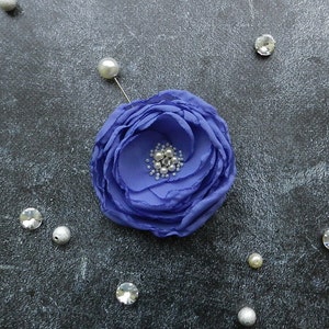 May include: A purple fabric flower brooch with a pin on the back. The flower has a center made of small white and silver beads.
