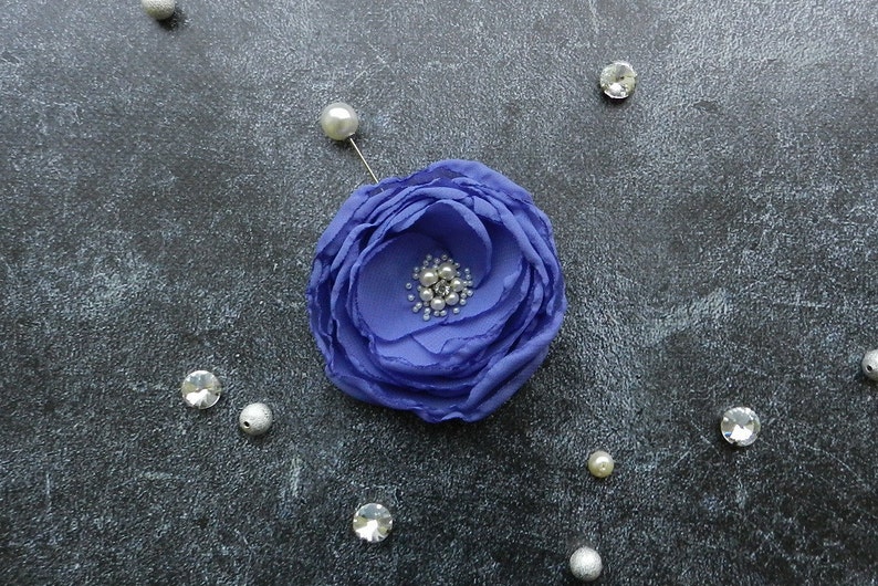 May include: A purple fabric flower brooch with a pin on the back. The flower has a center made of small white and silver beads.