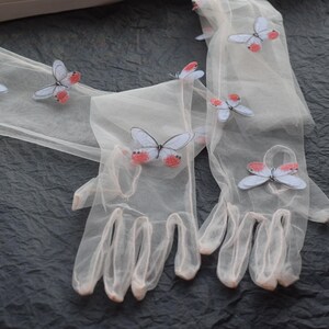 May include: Sheer, light-colored gloves adorned with butterfly appliqu&eacute;s. The butterflies have pale blue wings and red accents. The gloves are made of a delicate, translucent material, and are displayed on a dark, textured surface.