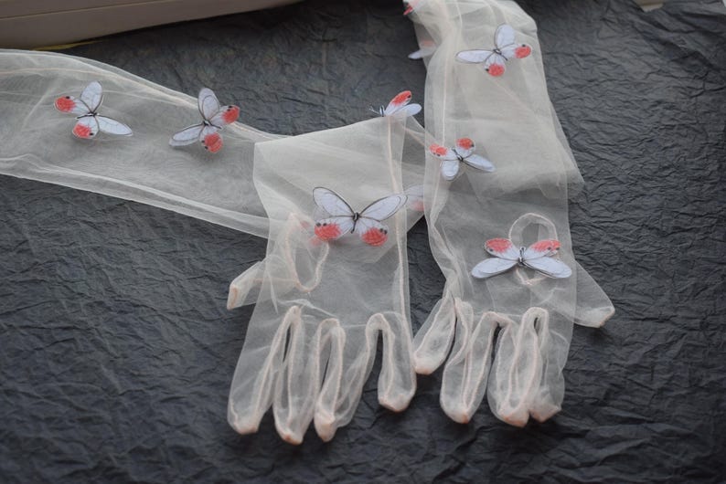 May include: Sheer, light-colored gloves adorned with butterfly appliqu&eacute;s. The butterflies have pale blue wings and red accents. The gloves are made of a delicate, translucent material, and are displayed on a dark, textured surface.