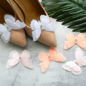 May include: A pair of beige shoes with white and pink butterfly embellishments. The butterflies have a pearl-like center and are layered for a delicate look.