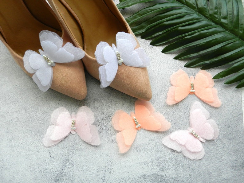 May include: A pair of beige shoes with white and pink butterfly embellishments. The butterflies have a pearl-like center and are layered for a delicate look.