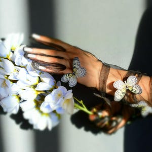 May include: A hand wearing a black mesh sleeve adorned with butterfly appliqués holds a bouquet of white flowers with yellow centers. The butterflies have white and gray patterned wings.