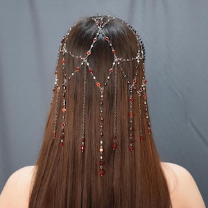 Puede incluir: Un adorno para la cabeza con una red de cadenas plateadas y cuentas rojas y blancas. El adorno para la cabeza se lleva en la parte posterior de la cabeza, con cadenas y cuentas cayendo en cascada por el cabello. Las cuentas son de varios tamaños y formas.