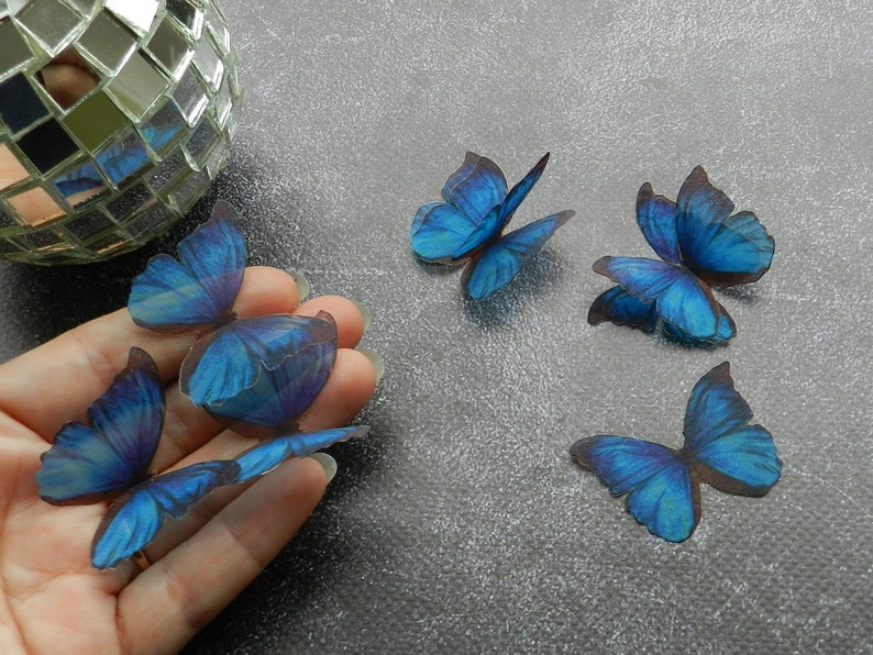 May include: Six blue butterfly shaped paper cutouts. The butterflies are arranged in a hand and on a gray surface.