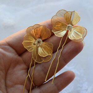 May include: Two gold-colored hair pins, each featuring a delicate, wire-framed flower design. The flowers have multiple layers of petals and a small, clear bead in the center. The pins are held in a hand against a soft, white background.