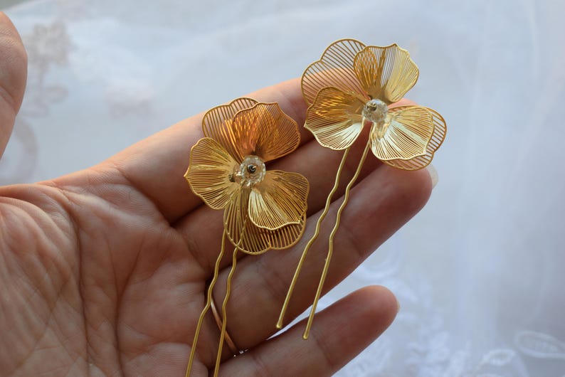 May include: Two gold-colored hair pins, each featuring a delicate, wire-framed flower design. The flowers have multiple layers of petals and a small, clear bead in the center. The pins are held in a hand against a soft, white background.