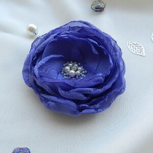 May include: A purple fabric flower brooch with a pearl center. The brooch is pinned to a white fabric background.