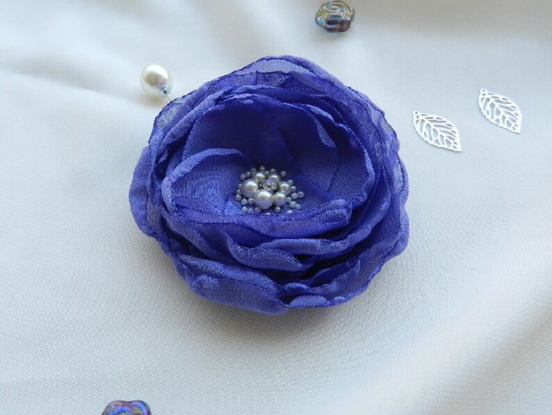 May include: A purple fabric flower brooch with a pearl center. The brooch is pinned to a white fabric background.