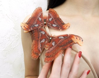 May include: Three large, brown and white butterfly wings with black accents are pinned to a person's shoulder. The wings are made of fabric and have a realistic appearance.